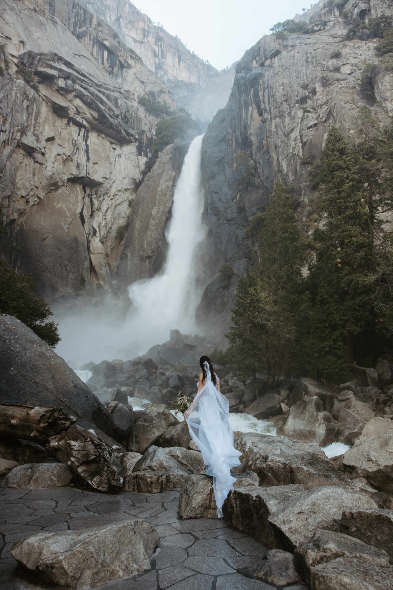 Bride standing beneath Yosemite Falls in her wedding dress, feeling the mist during her intimate Yosemite elopement.